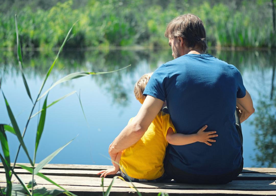 Parent and child hugging sitting on wooden dock on a lake.