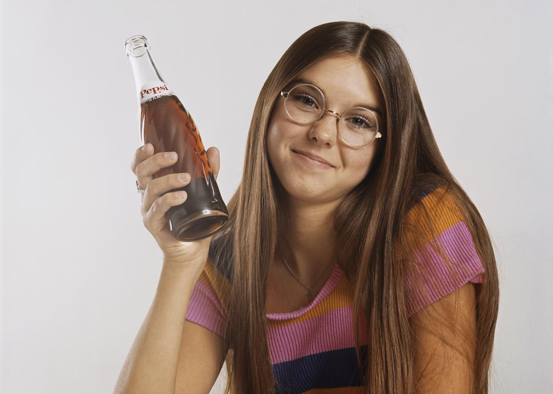 A teenager with long hair and glasses in the 1970s holding a soda in a glass bottle.