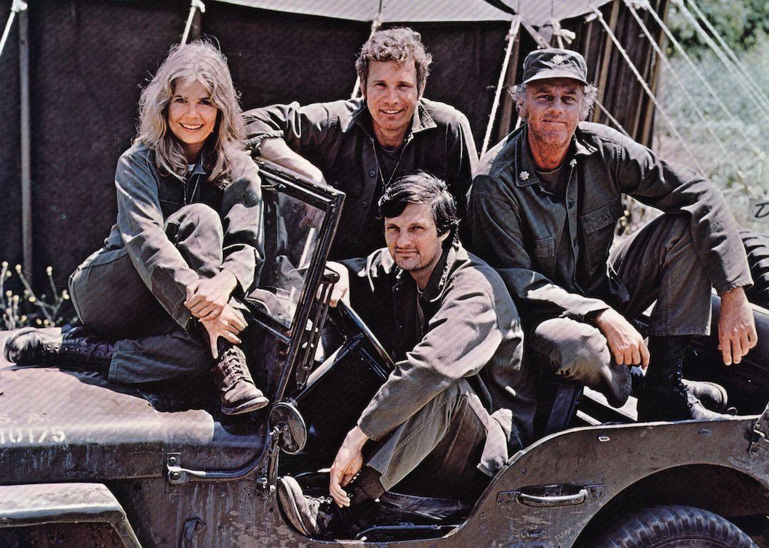 Alan Alda in the driving seat of a jeep, surrounded by Loretta Swit and other cast members of the hit television show 'M*A*S*H.'