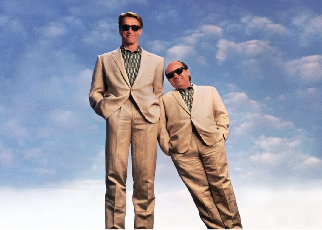 Danny DeVito leans on Arnold Schwarzenegger in matching suits in the movie 'Twins.'
