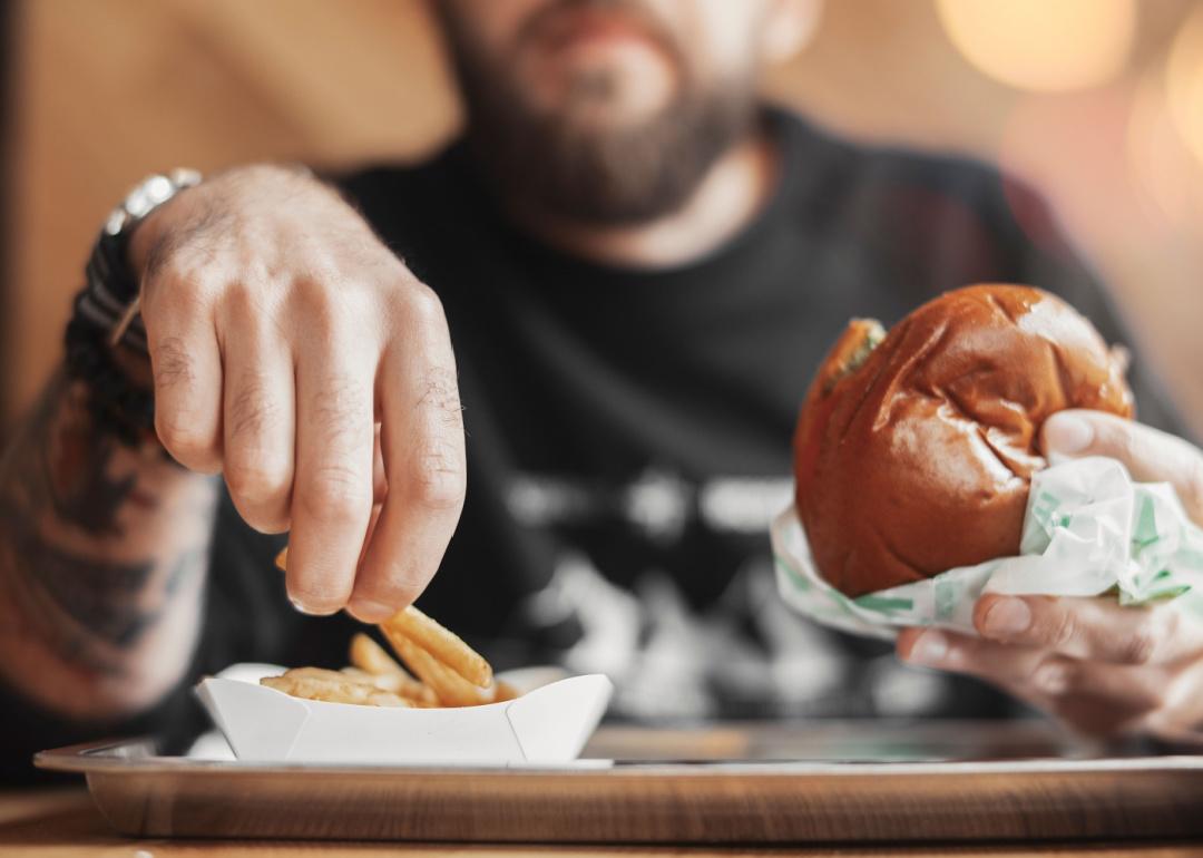 A diner picks up a fast-food french fry with one hand and holds a hamburger in the other.
