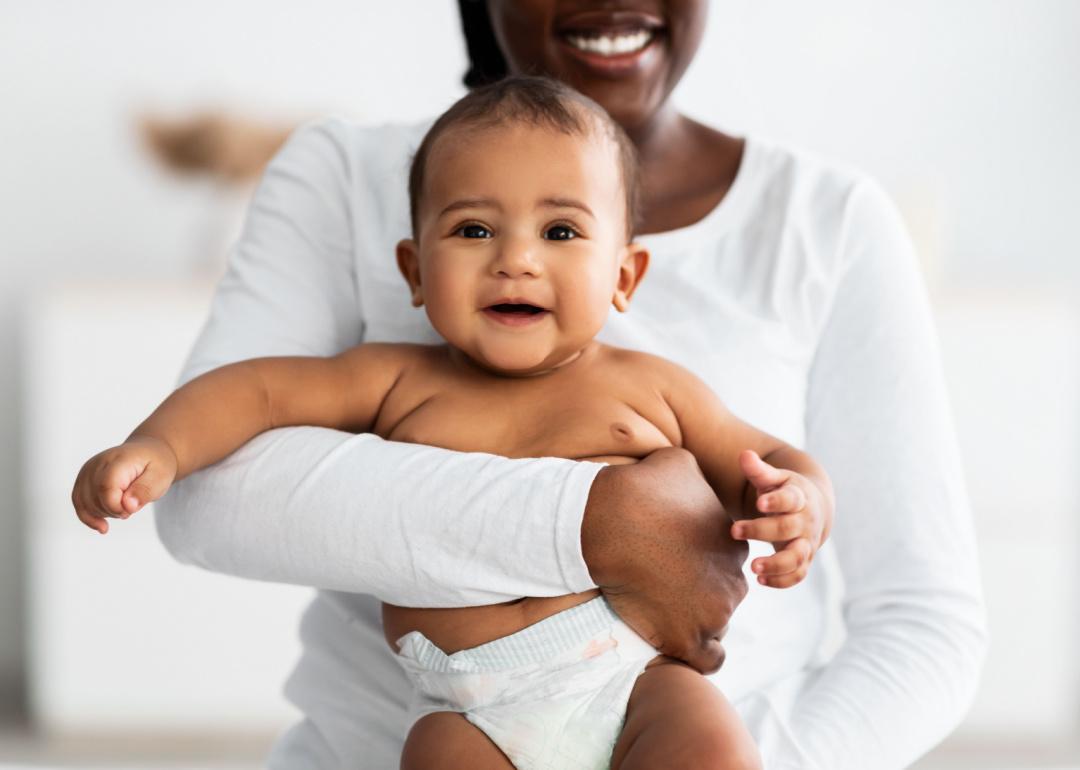 Parent wearing a white long-sleeved shirt holds smiling newborn baby in a diaper. 
