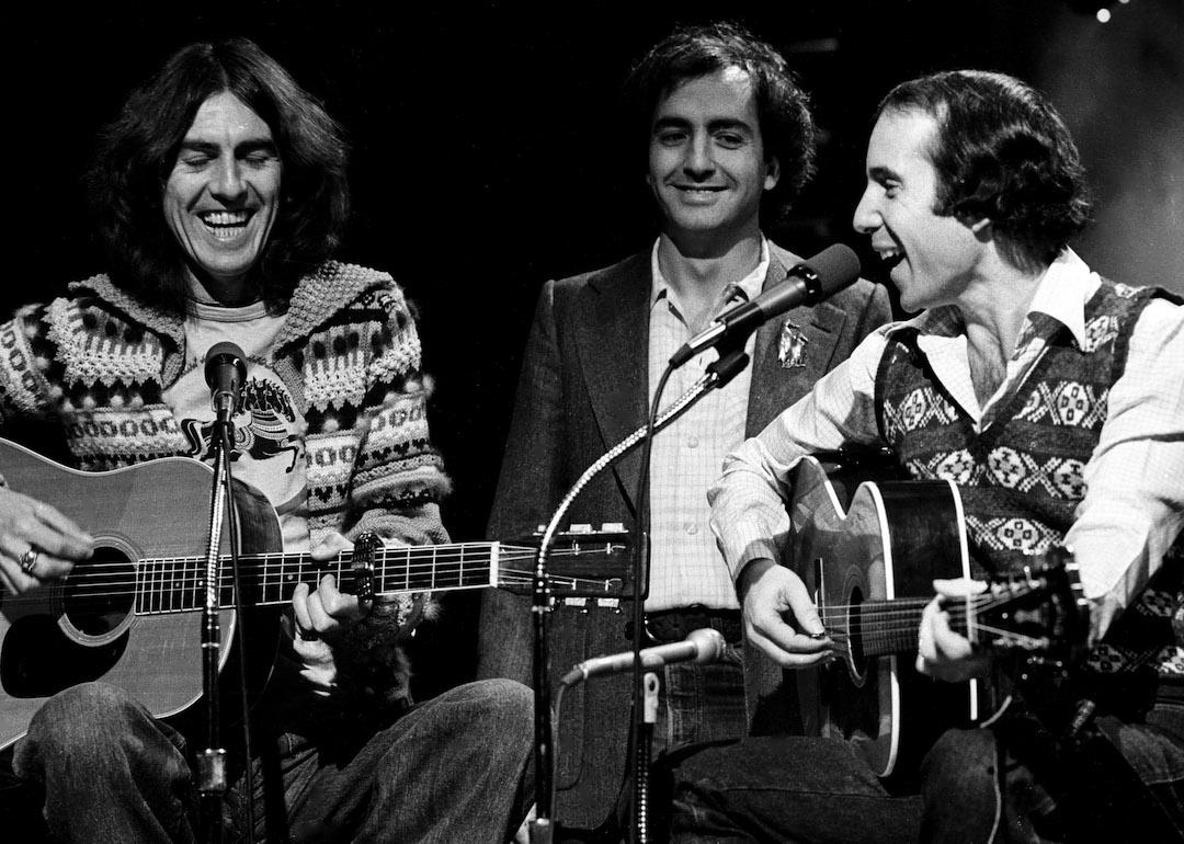 Lorne Michaels stands behind George Harrison and Paul Simon as they perform on 'SNL' in 1976.