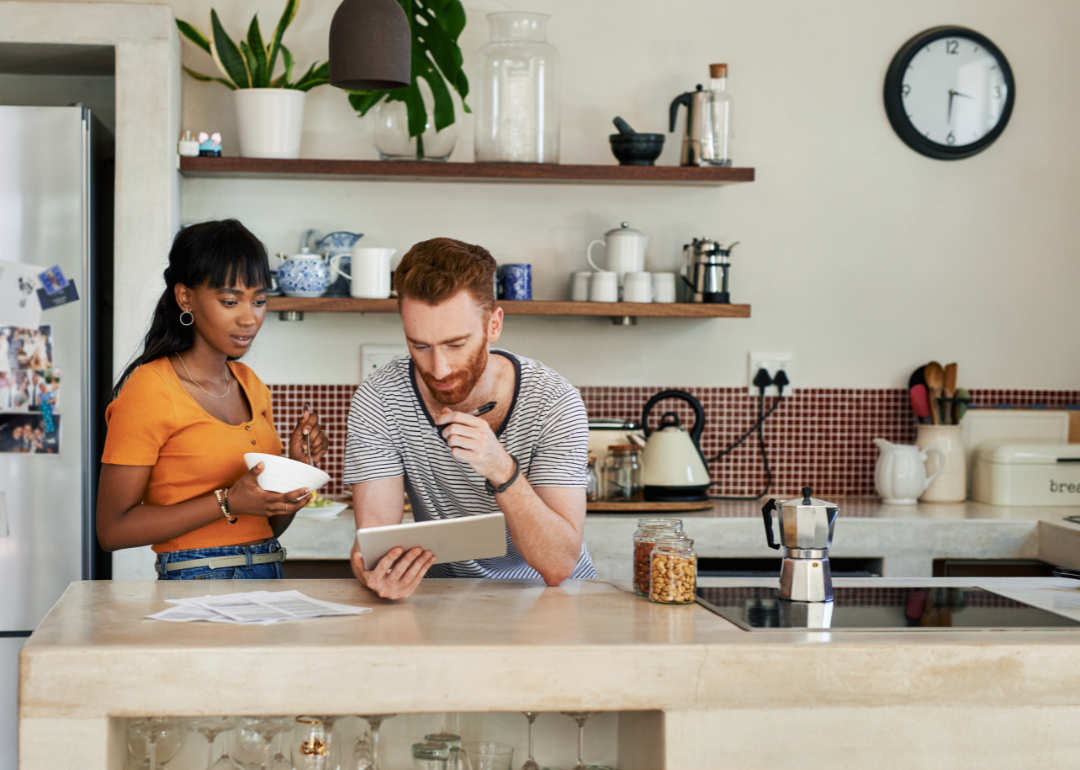 A young couple jointly study a laptop in a kitchen setting.