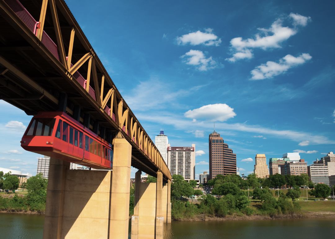 A suspended red tram moves beneath a yellow bridge with the Memphis skyline in the distance.