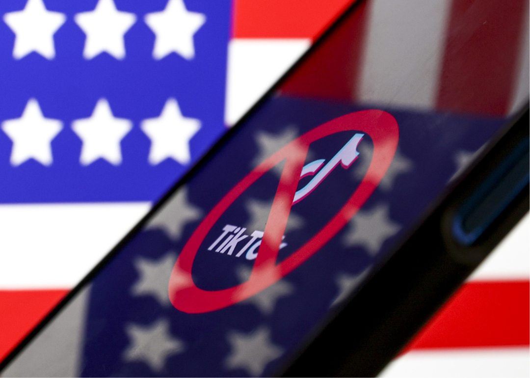 Image shows a mobile phone with the tiktok logo on the screen that has a red X through it. American flag is blurred in the background.