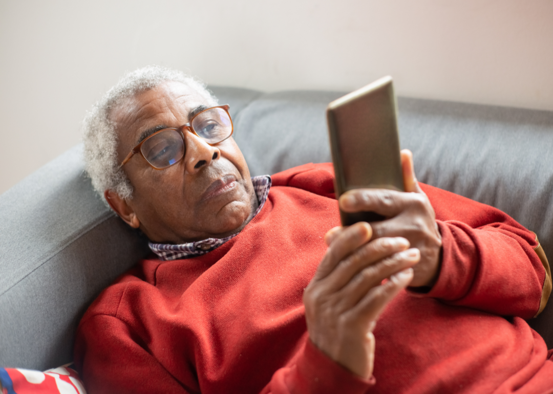Senior with white hair wearing a red sweater lying on the couch reading a message on their smart phone.