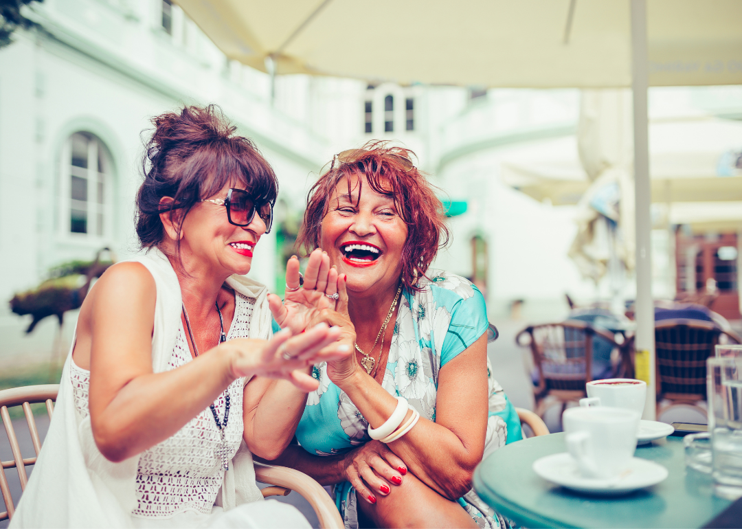 Two senior friends laugh together while sitting at a cafe in a city.