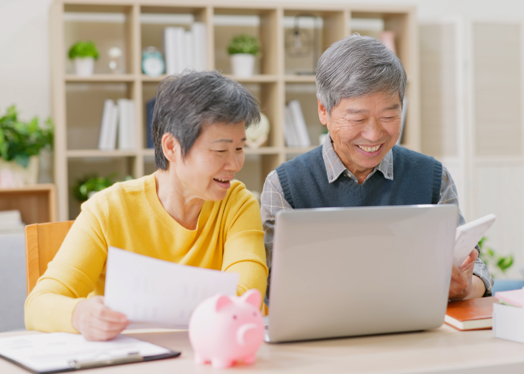 An elderly couple looks at a computer, smiling, with a small piggy bank in front of them.