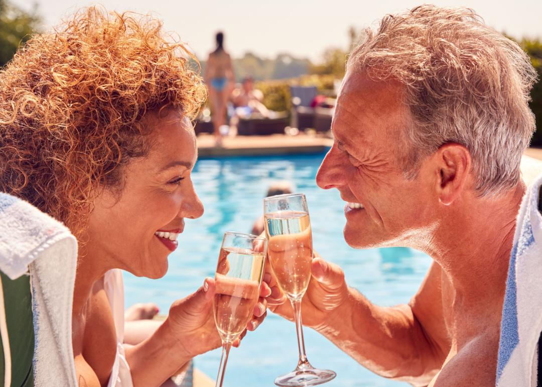 A senior couple toasts champagne while sitting by the pool.