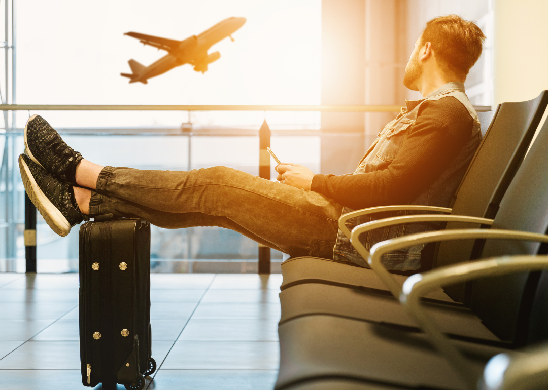 A man sits in a chair at an airport with his feet up on his carryon suitcase, watching a plane take off in the distance