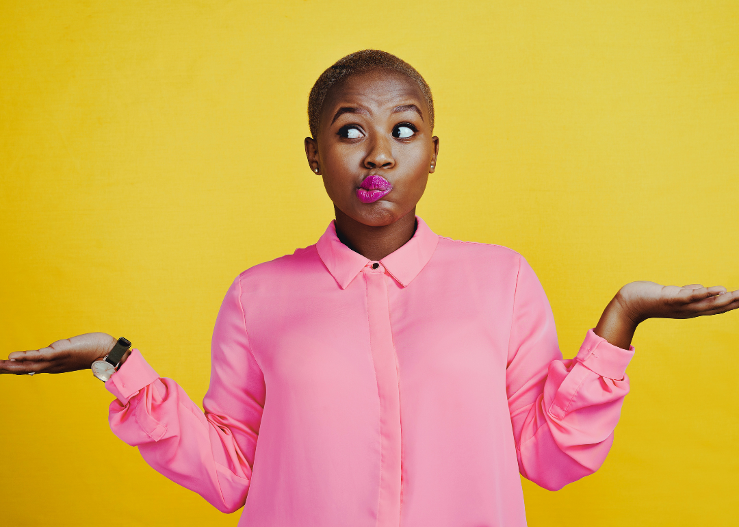 A Black woman in a pink long-sleeved blouse holds up both hands in a gesture of indecisiveness.
