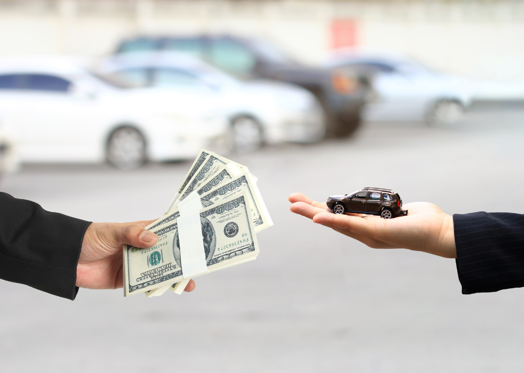 A conceptual image showing a financial transaction: one hand holding a small model car and another hand holding a stack of $100 dollar bills