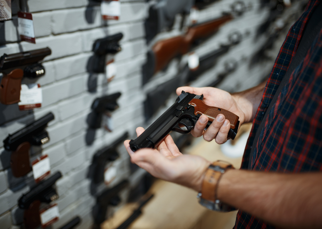 Hands holding and checking out a hand gun in gun shop, a wall of guns in the background