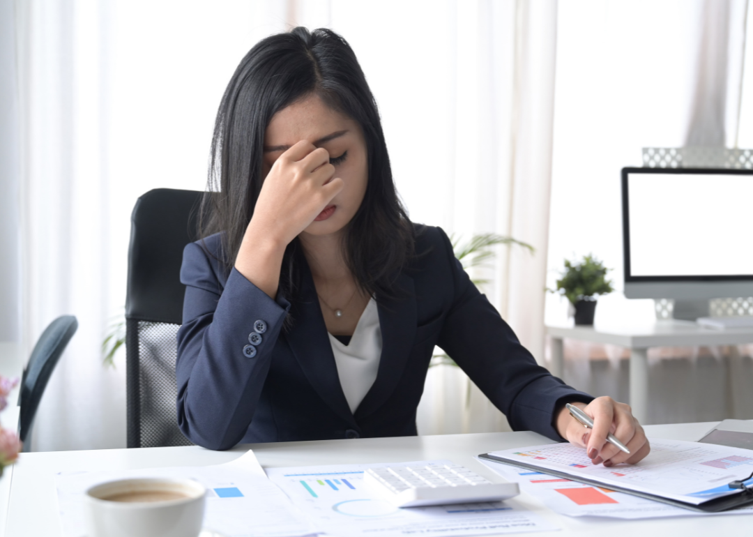 A businesswoman seated at a desk holds a hand to her face in worry.