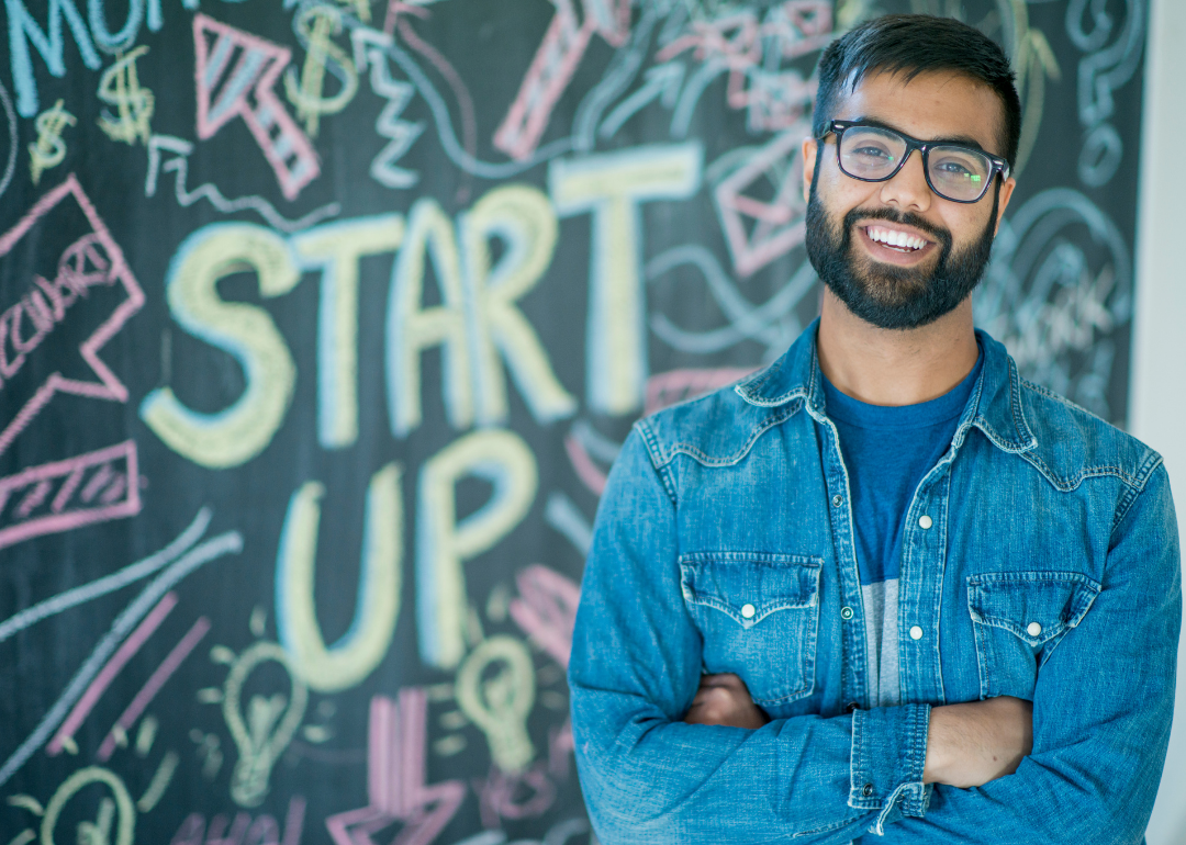 A young Indian man with a beard and glasses, smiling, stands in front of a sign reading "START UP"