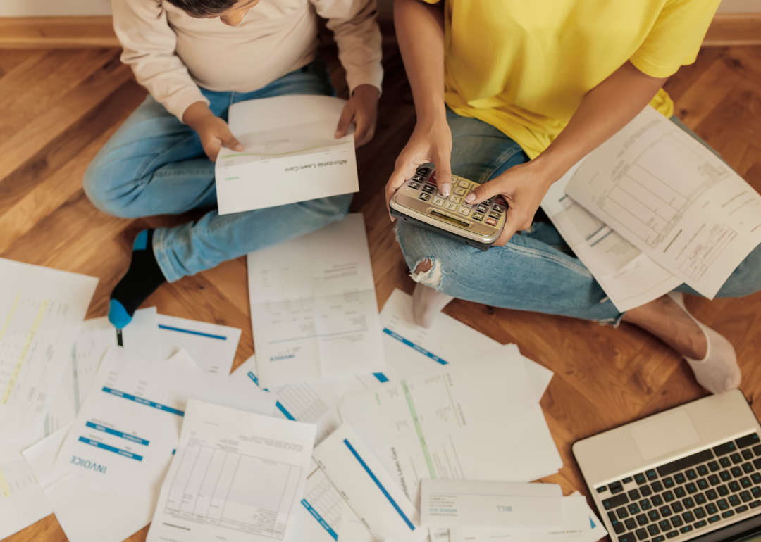 Two people sit on the floor amidst a scattering of paper bills and invoices, one of them using a calculator as they pay bills