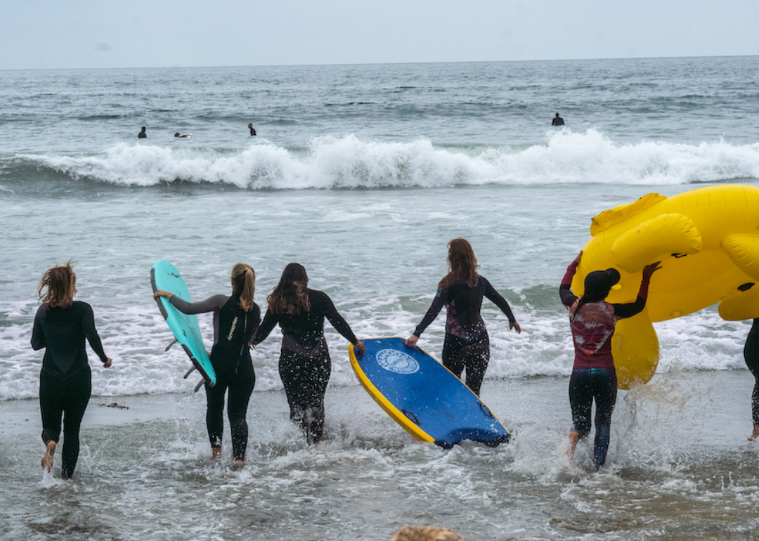 A group of women in surf wear run into the ocean with body boards and an inflated floatie