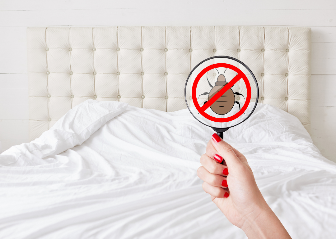 A woman's manicured hand holds a magnifying glass over a hotel room bed to check for bed bugs
