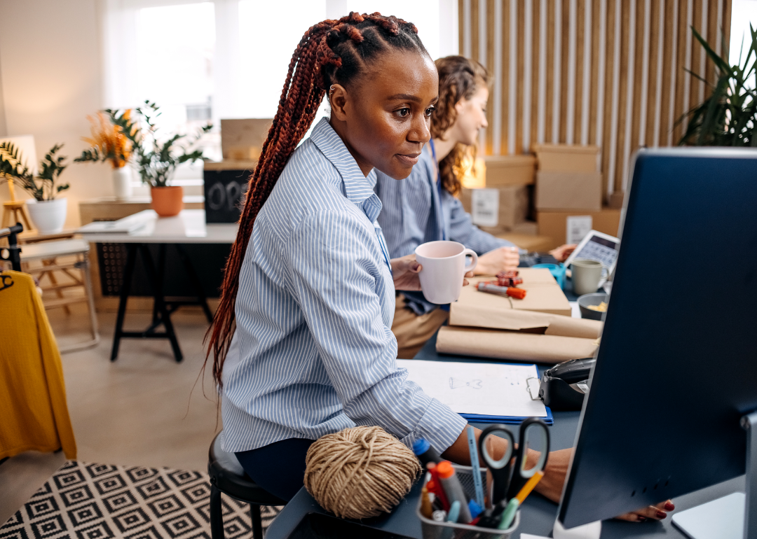 Two women sitting in a creative-looking office with plants and boxes, working on the computer and wrapping something in brown paper.