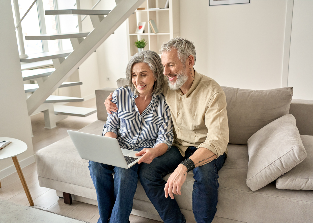 A senior couple sit on a beige couch and look at a laptop computer