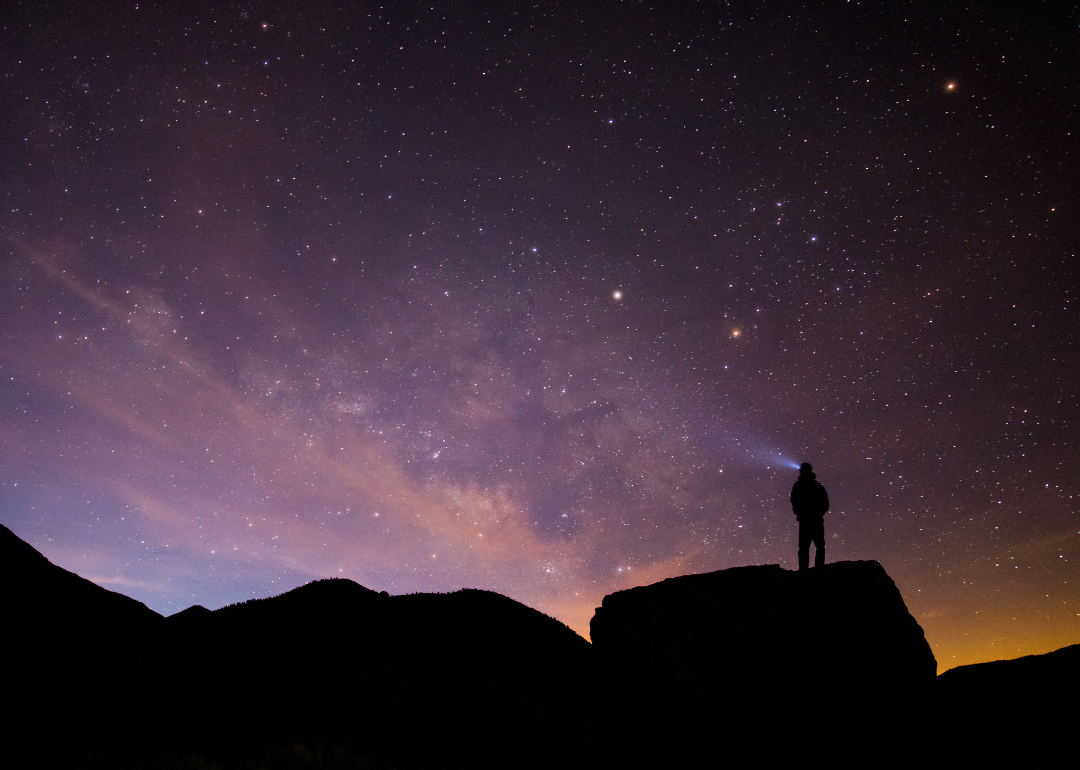 Silhouetted person stands atop boulder with light beam looking up at the milky way and sunrise.