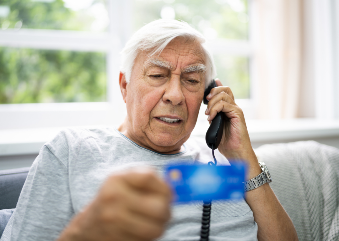 An older man is on the phone, looking distressed, holding up his credit card