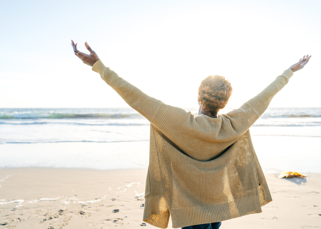 An older woman in a cardigan sweater stands on a beach with her arms raised