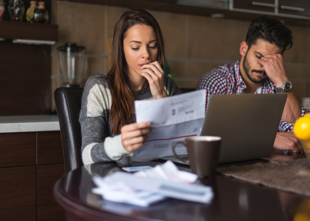 A young couple sit at a table. On the table is a laptop. The woman is holding a bill in her hands and looks worried. The man has has head in his hands.