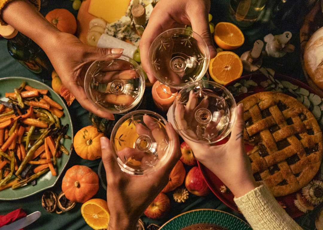 Four people toast with wine glasses over a Thanksgiving meal. Only their hands and the glasses are shown above the table.