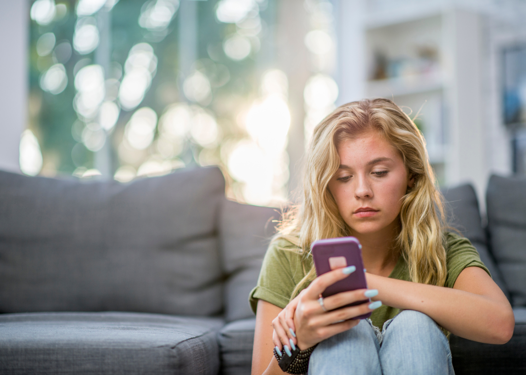 A young blonde girl sits in front of a gray couch and looks at her smartphone