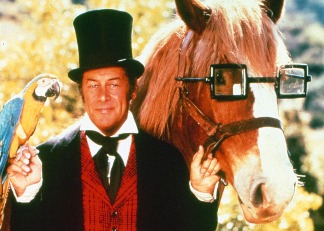 Actor Rex Harrison wearing a long black coat, a red waistcoat, and a black top hat, with a parrot sitting on his right hand as he stands beside a horse wearing a pair of glasses in a publicity portrait for the 1967 film 'Doctor Dolittle.'