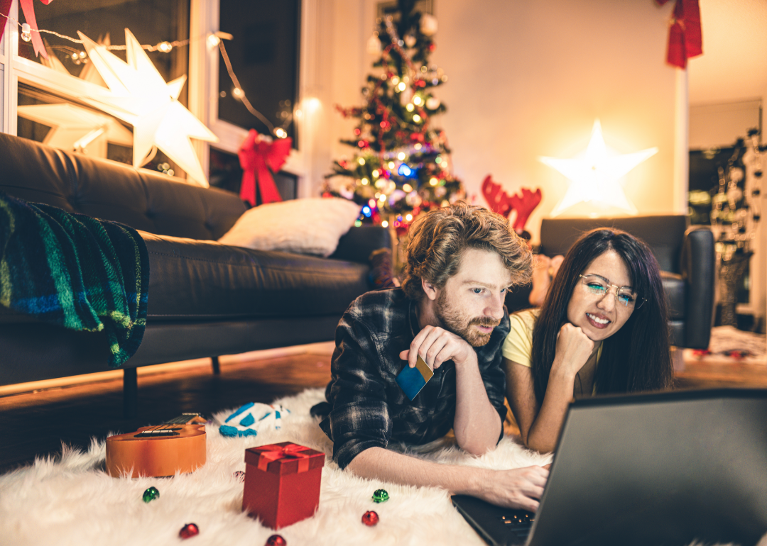 A couple lie on the carpet in their sitting room and shop online from a laptop; a Christmas tree is lit in the background