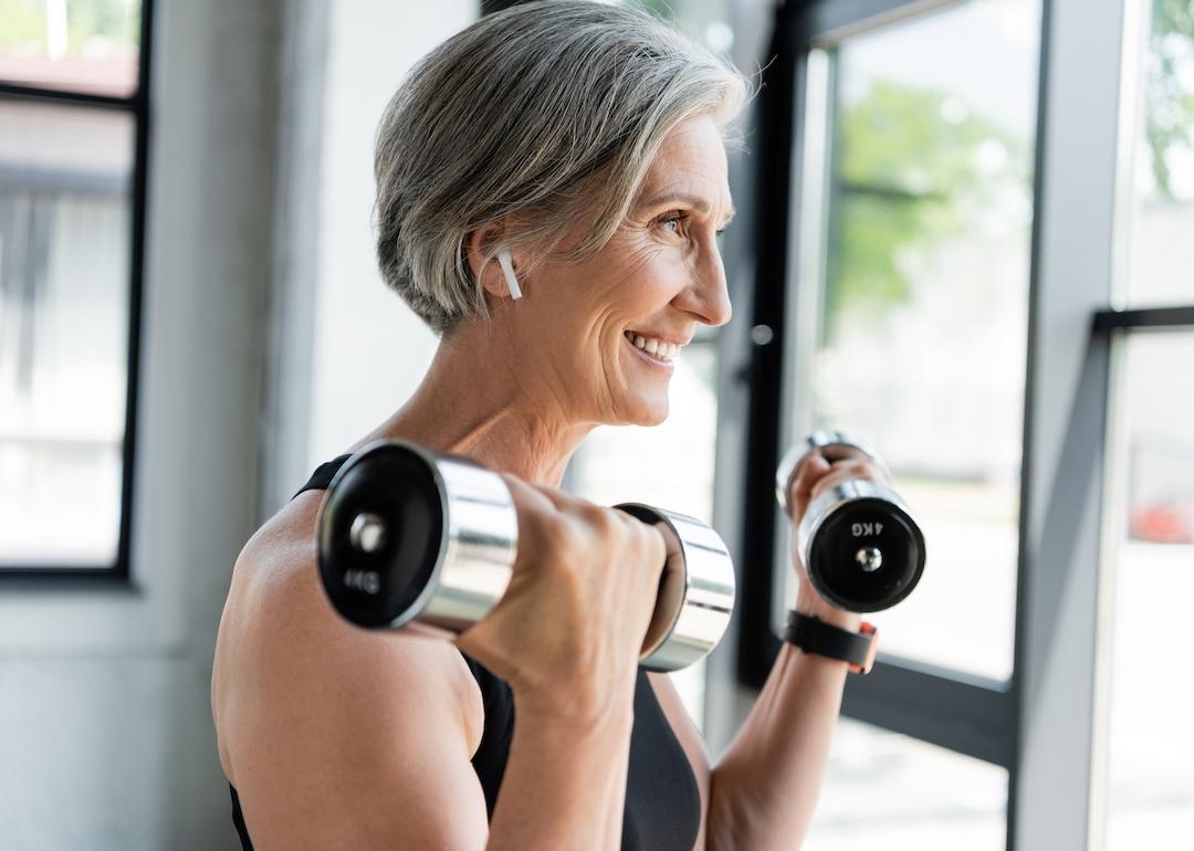 Smiling person with gray hair lifts weights while wearing wireless headphones.