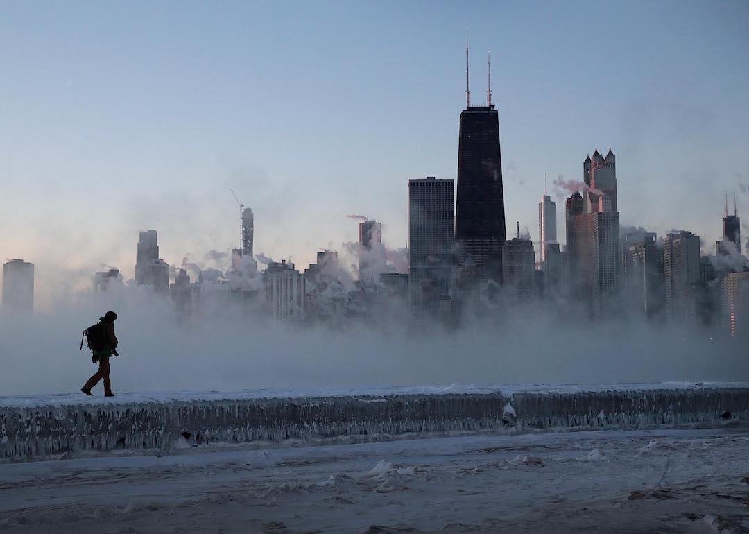 A person in a parka walks along an icy surface with the Chicago skyline in the background on a cold, windy day.
