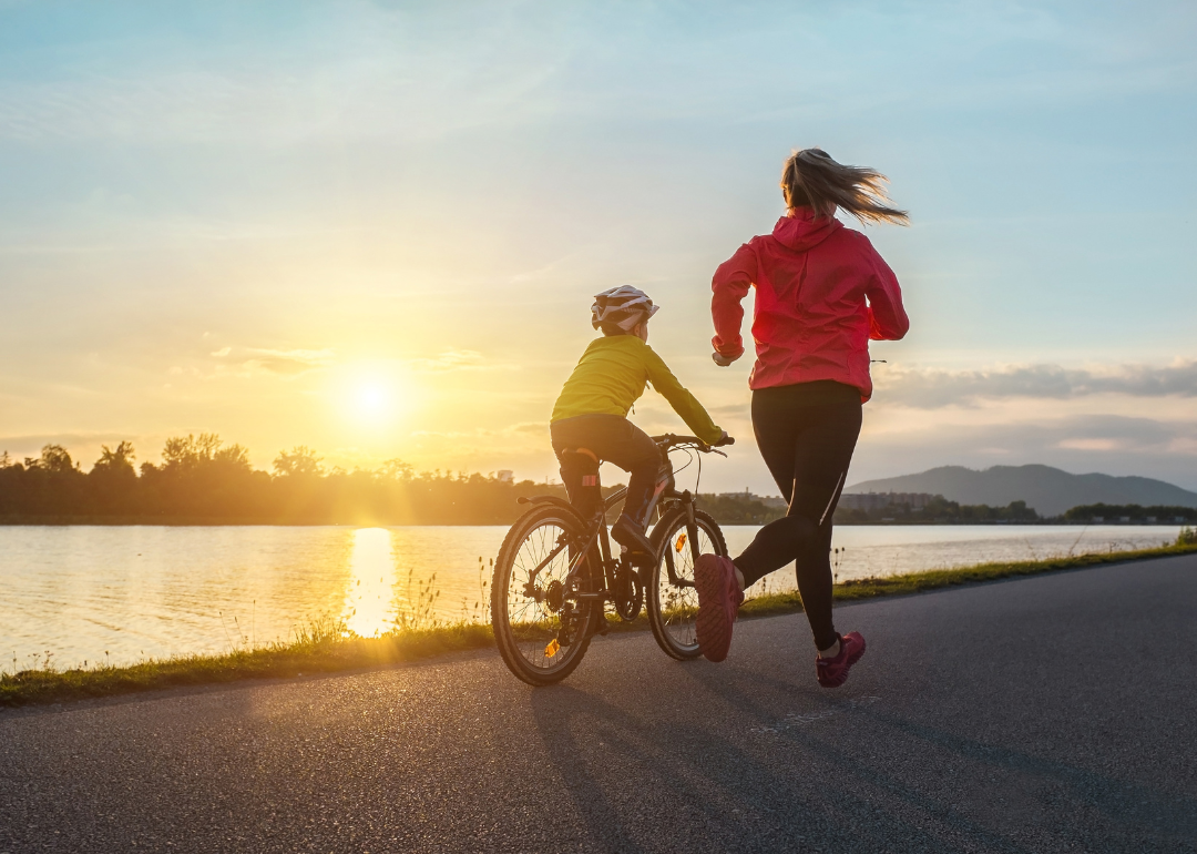 Mother running alongside her son on a bicycle on a waterfront path at sunrise or sunset