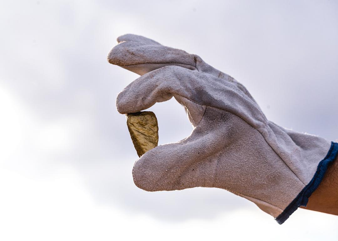 Gloved hand holding up a gold nugget between their pointer finger and thumb.
