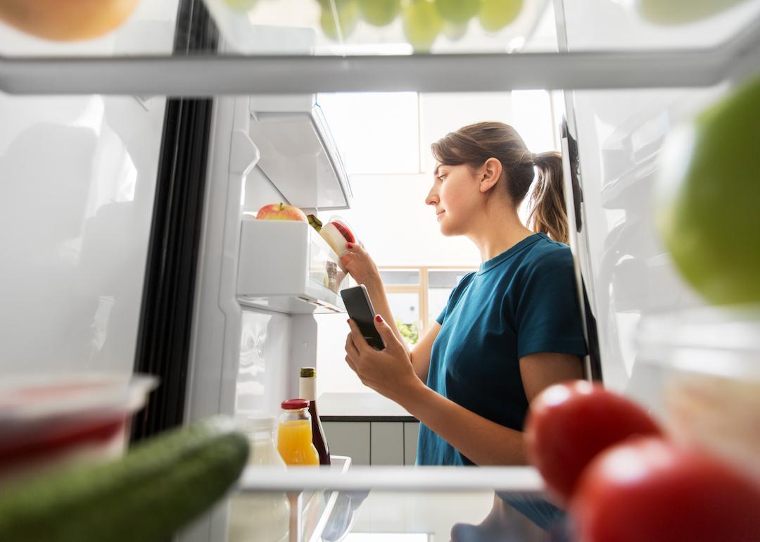Person looking at the food in the door of their fridge with a smartphone in their hand.