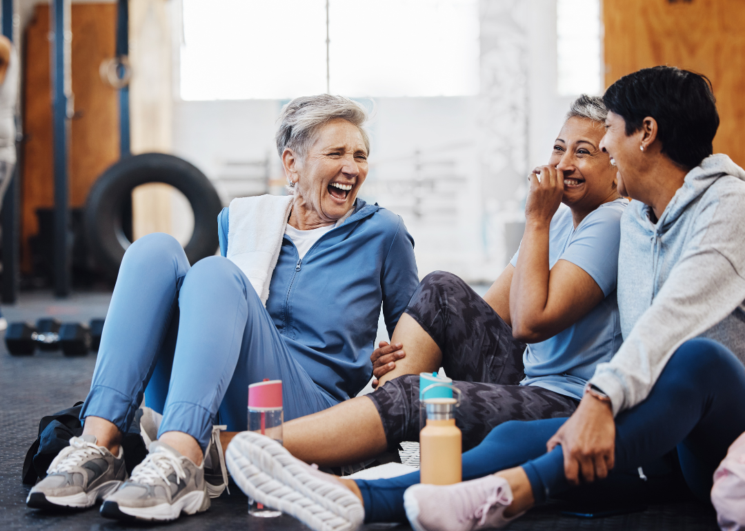 Three older women sitting on a gym floor and laughing