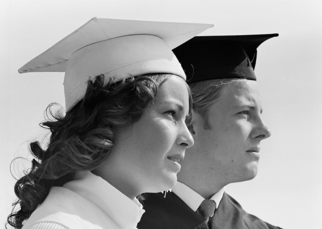 Close-up of two college graduates wearing a white and black cap and gown respectively and looking off into the distance, circa 1970s