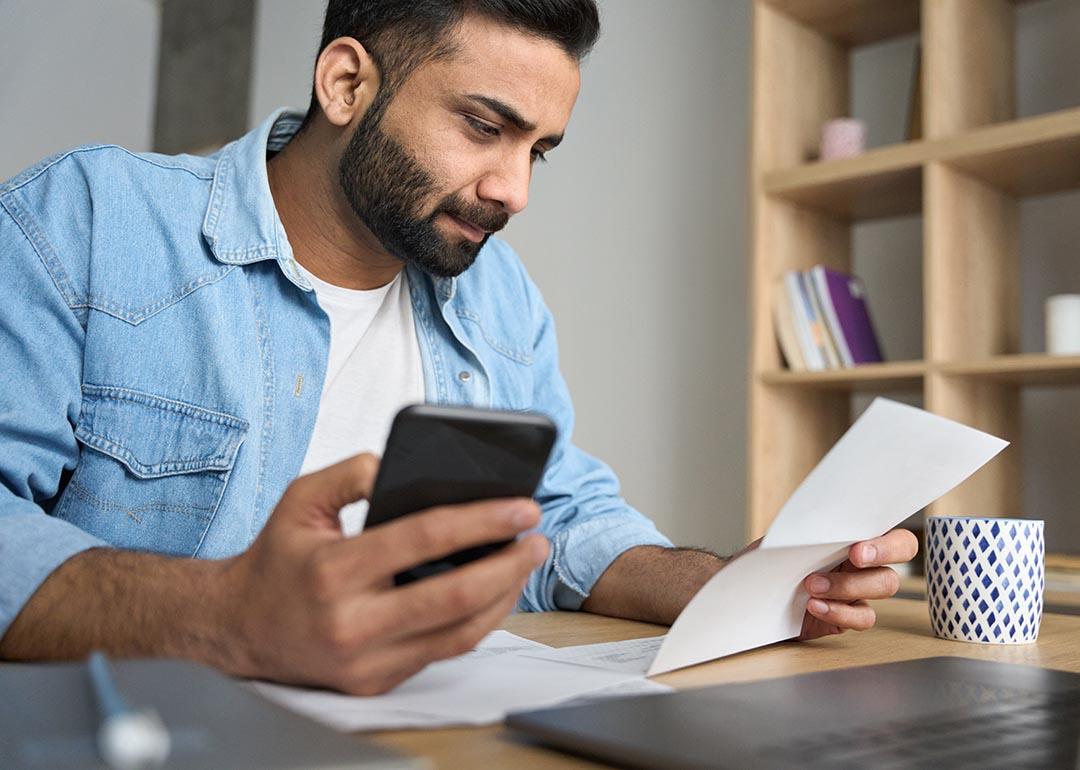 A college student seemingly calculating bills holding a phone with one hand and holding a paper bill with the other.