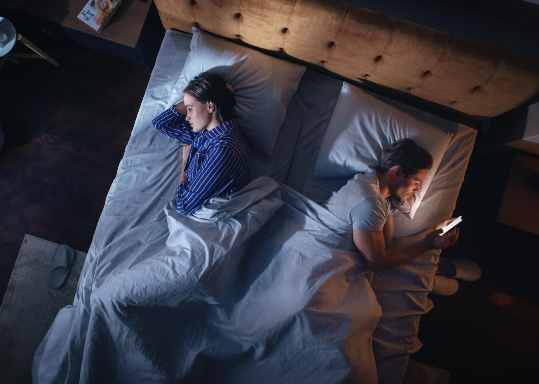 Overhead view of couple laying in bed facing away from each other in the dark while one person is scrolling on phone.