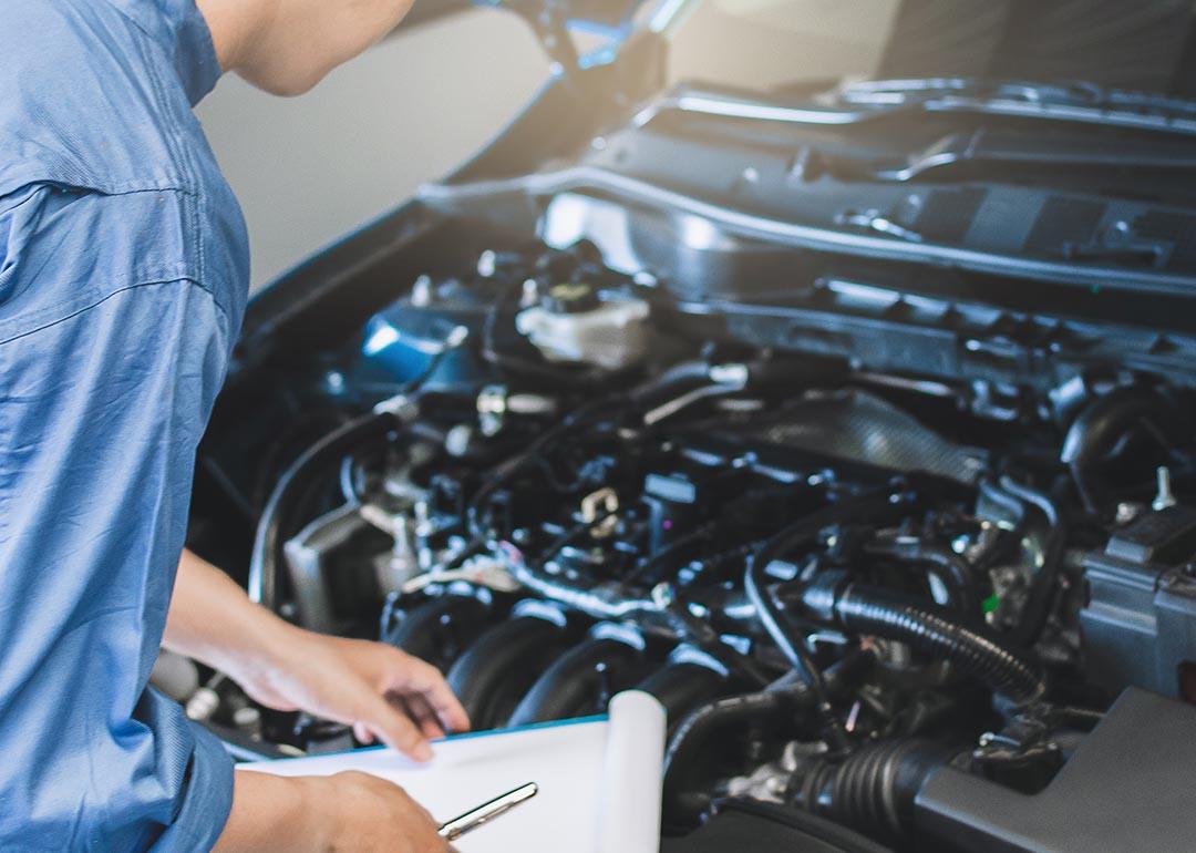 Mechanic in blue suit with clipboard inspects under the hood of car.