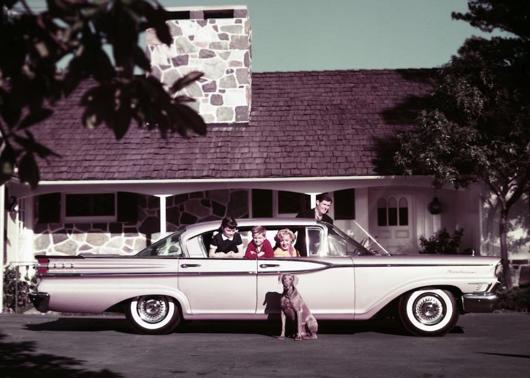 A family of four in their Mercury Monterey automobile in the driveway of their home with their pet dog sitting next to the car.