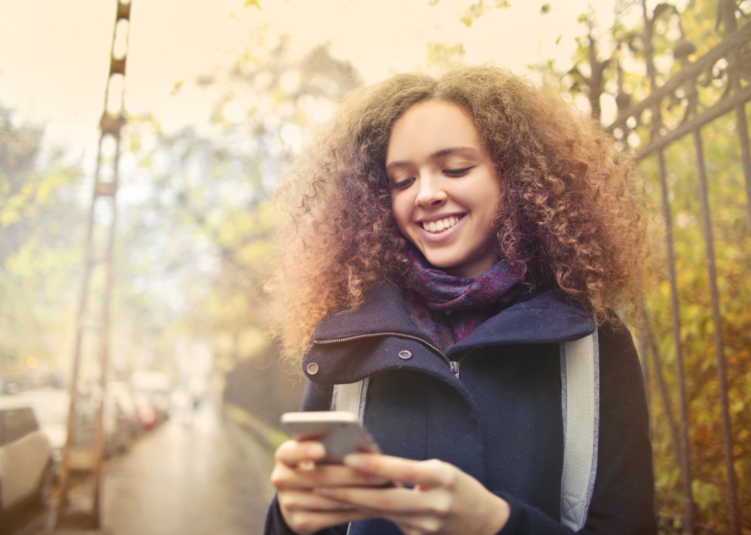 A smiling young woman looks at her smartphone while outside.