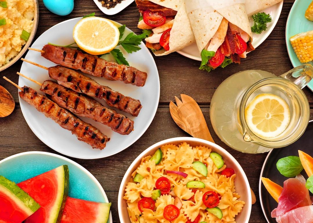 Summer food on wooden table, including pasta salad and BBQ grilled skewers.