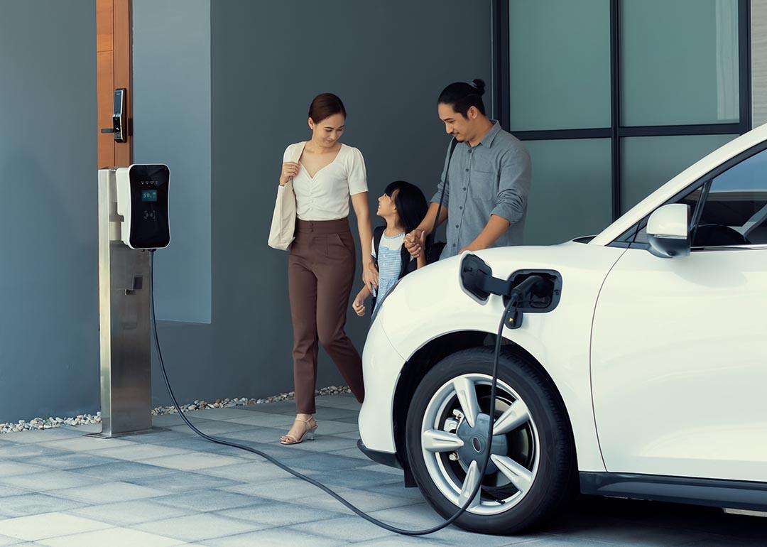 A family of three happily walking towards their electric car being charged in a station.