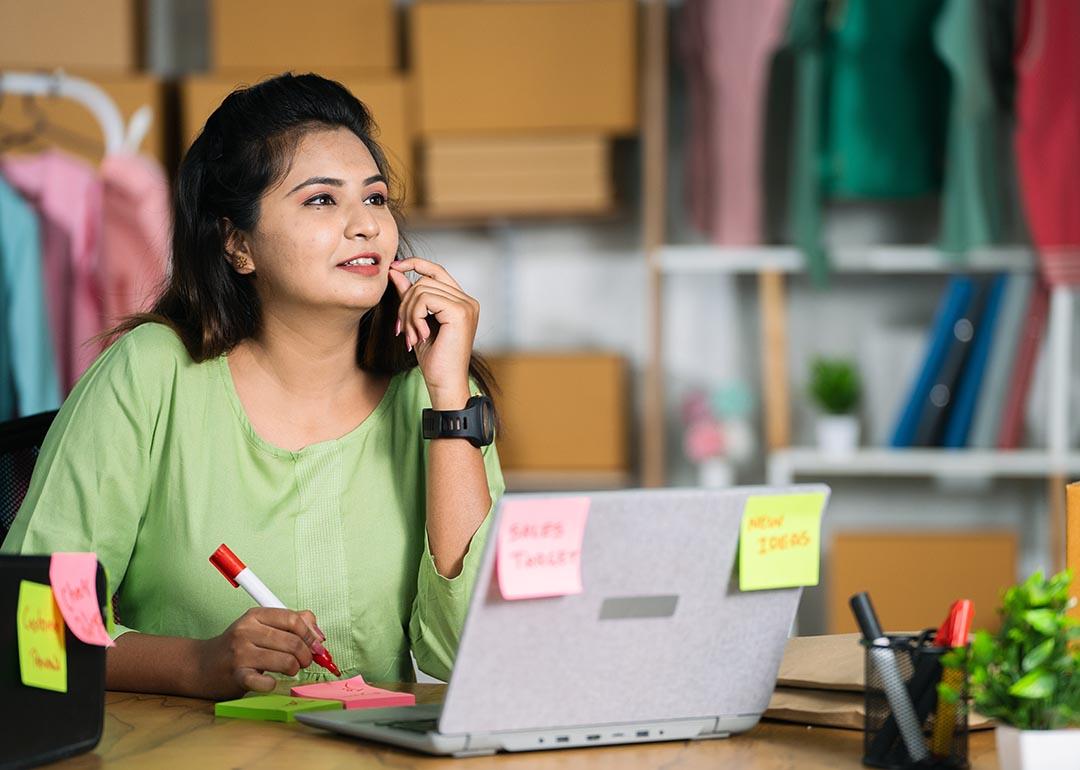 A woman inside her small business studio is seemingly contemplating about ideas using a laptop and while writing on sticky notes.