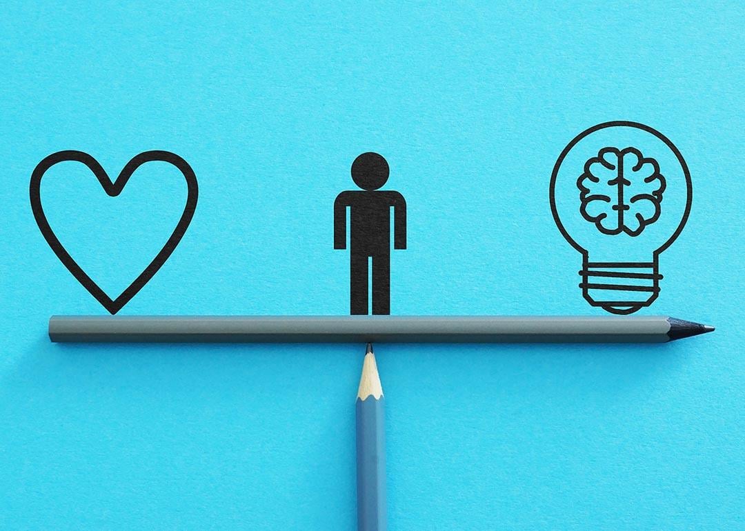 A minimalist illustration on a cyan background. Two pencils resemble a weighing scale with a human figure in the middle, a heart symbol on the left side, and a bulb with a brain on the right.