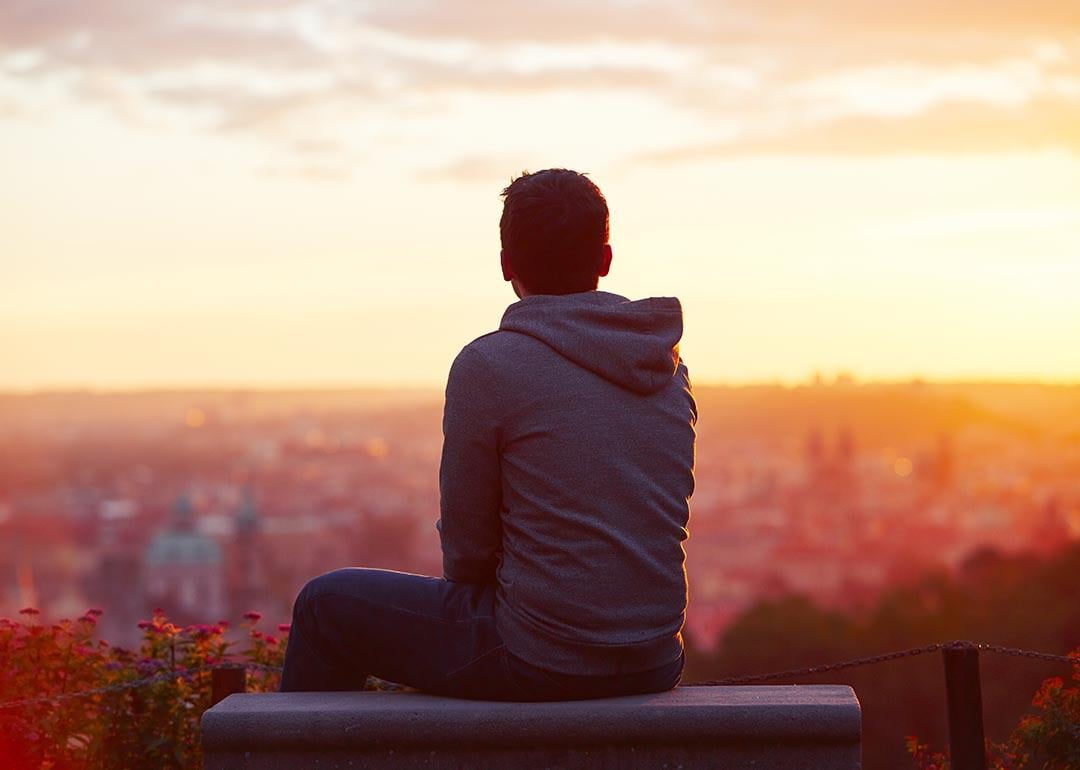 A man is sitting on a bench overlooking the city during sunset.